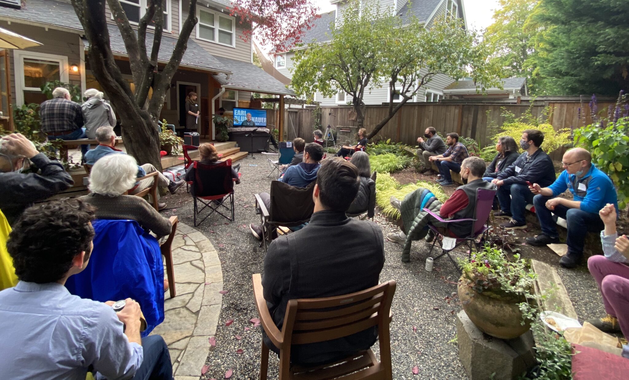 Photo of a Parking Reform Network event in a backyard. People are sitting around on chairs watching a video address by Earl Blumenauer.
